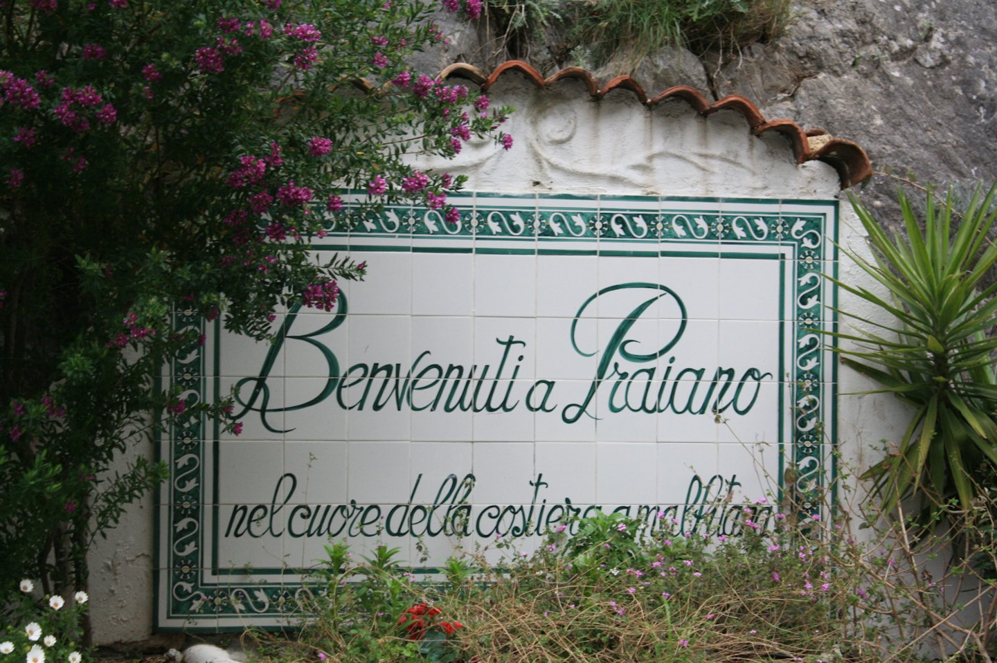 Cartello in ceramica di benvenuto a Praiano su un muro intonacato.